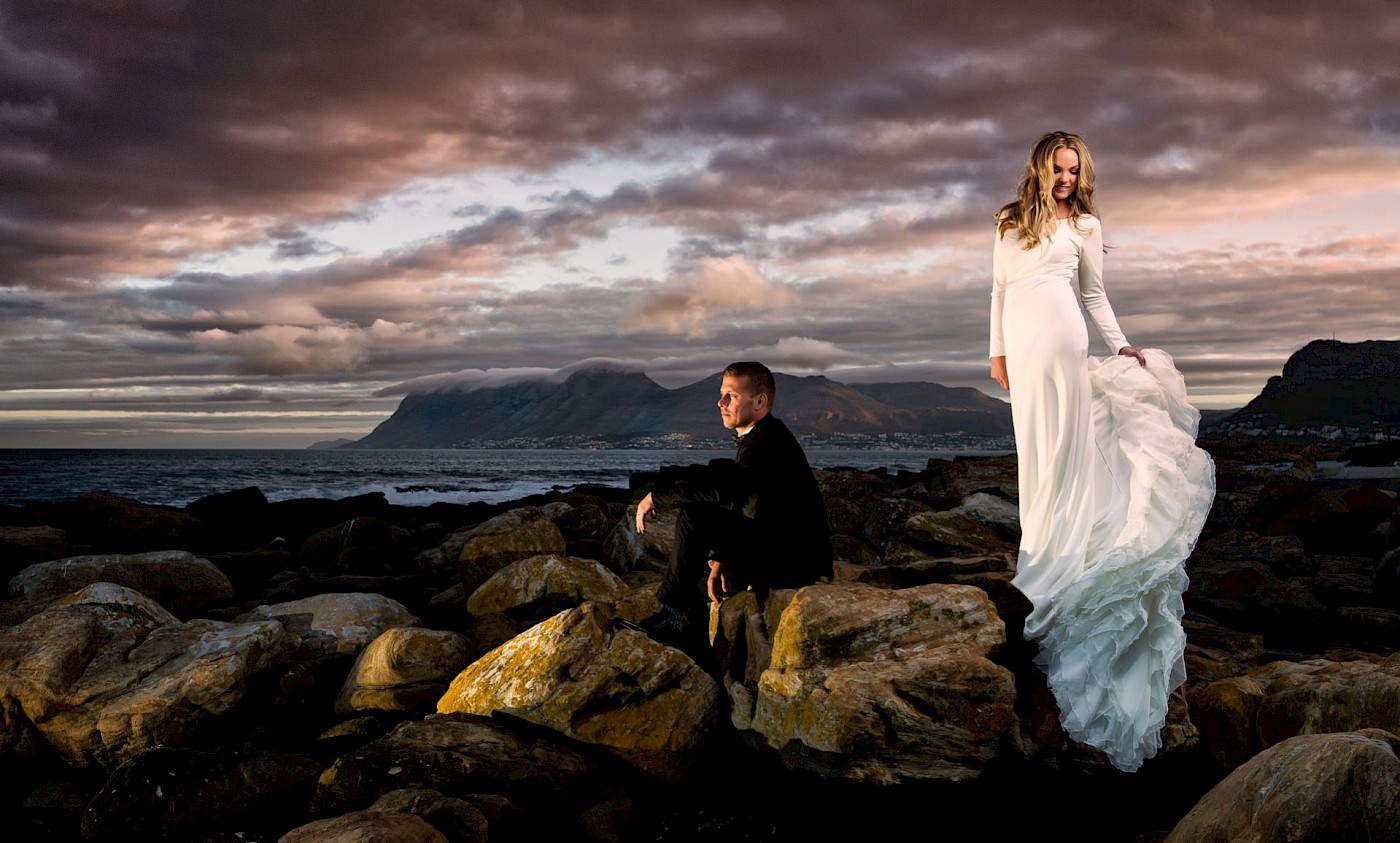 Stunning dramatic image of newlyweds on a rocky beach - him sitting in a tux - her waving her dress - photo Greg Lumley