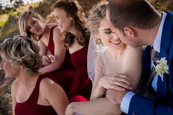 A bride and groom share an affectionate moment while the rest of the wedding party chats and laughs in the background – Photo by Greg Lumley