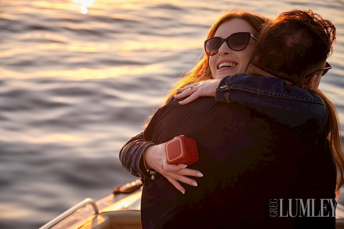 A joyful red-haired woman embraces her fiancé after getting engaged, both smiling on a yacht with the ocean in the background.