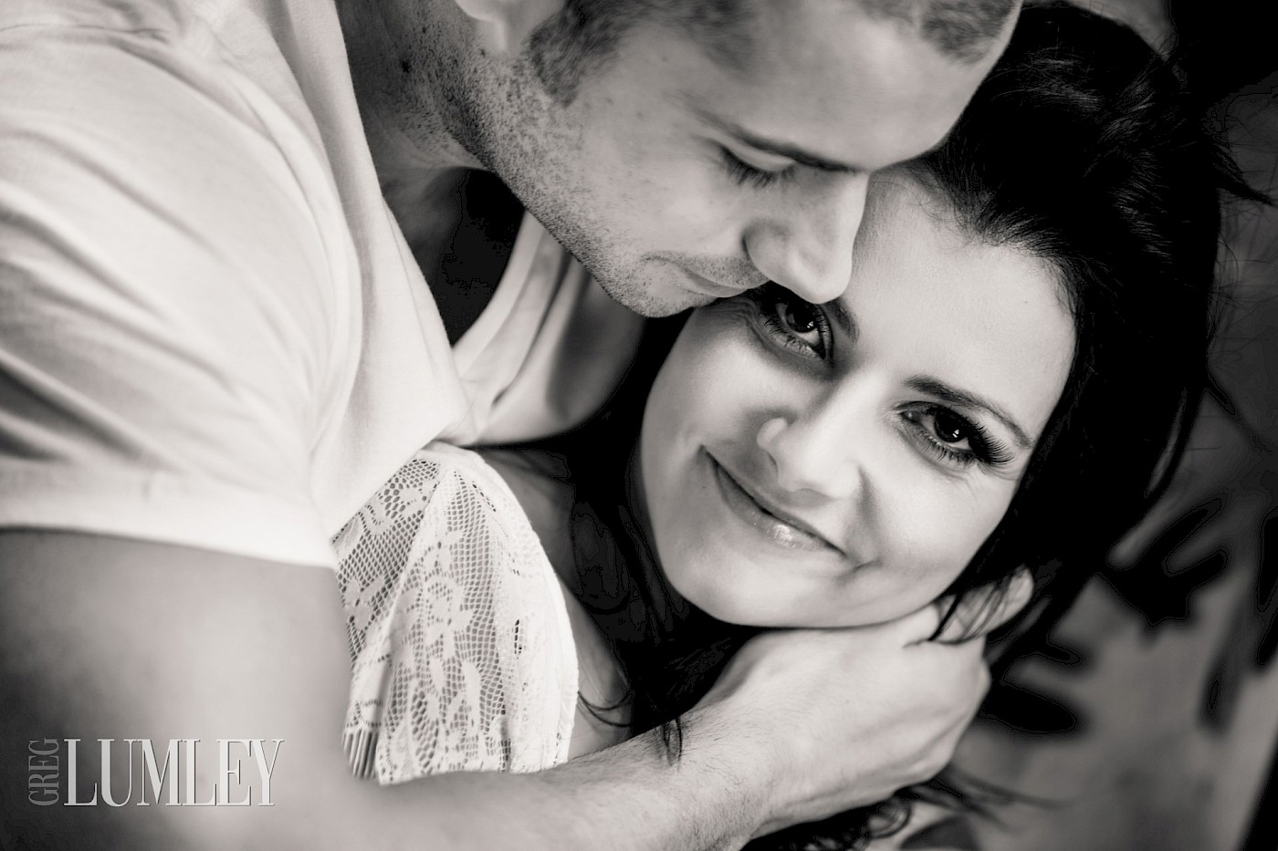 A young man gently holds his fiancée close. She looks straight into the camera, smiling with quiet joy. A timeless black and white moment filled with warmth.